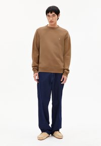 Jonge man in een bruine sweatshirt, losse donkerblauwe jeans en beige mocassin schoenen, staande tegen een effen witte achtergrond.