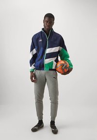 Mladic u šarenom sportskom sakou i sivim Puma trenirkama drži narandžastu i crnu fudbalsku loptu, stojeći na jednobojnoj pozadini.