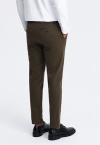Chino marroni con vestibilità su misura e dettagli sulle tasche posteriori, abbinati a scarpe eleganti nere e a una camicia bianca a maniche lunghe.