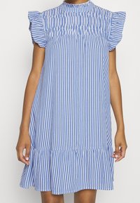 Robe à rayures bleues et blanches avec un col montant, un corsage froncé et des manches à volants. Longueur genoux avec un ourlet évasé. Tissu en coton.