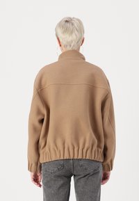 Giacca in pile marrone con colletto alto e orlo elasticizzato. Presenta spalle scese e maniche lunghe. Tessuto liscio e strutturato.