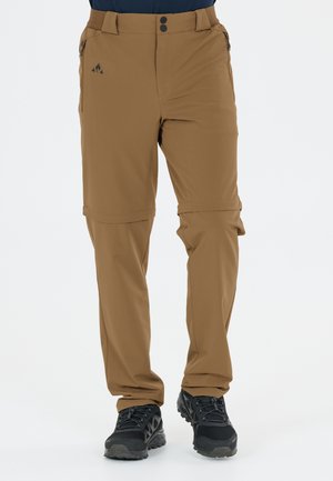 Braune Outdoor-Hose mit abnehmbaren Beinen, schwarze Wanderschuhe und ein in die Hose gestecktes dunkelblaues Hemd an einer stehenden Person.