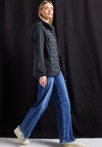 Schwarze Puffjacke mit Kapuze und Reißverschlusstaschen, getragen über blauen weit geschnittenen Jeans und weißen Sneakern. Glatte Textur, lässige Passform.