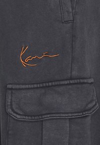 Mörkgrått tyg med orange broderad "Kani"-logotyp ovanför en stor ficka med klaff och synlig sömnad.