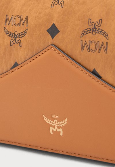 Surface en cuir fauve avec des logos MCM noirs répétés et des formes losanges, avec une pièce en cuir fauve lisse cousue, ornée d'un logo MCM doré.