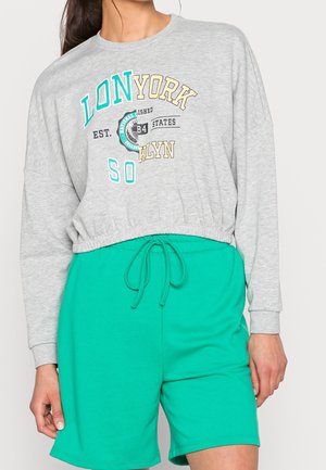 Avskuren grå sweatshirt med tryckt text i turkos, gult och svart; kombinerad med gröna shorts med dragsko. Mjuk, blandad bomullstextil.