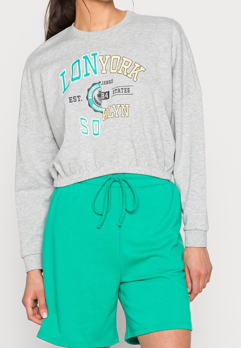 Sudadera gris recortada con texto impreso en turquesa, amarillo y negro; combinada con pantalones cortos verdes con cordón. Tejido suave de mezcla de algodón.