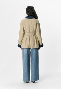 Beige gebelt jasje met een marineblauwe kraag en brede mouwen, gecombineerd met losse lichtblauwe jeans. Gladde textuur met een maatwerk ontwerp.