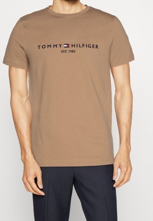 Mand iført en brun T-shirt med korte ærmer med teksten "Tommy Hilfiger EST. 1985" og logo, kombineret med mørke bukser.