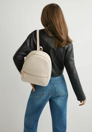 Kobieta o brązowych włosach, ubrana w czarną skórzaną kurtkę i niebieskie dżinsy, niesie beżowy skórzany plecak, stoi tyłem na jednolitym tle.