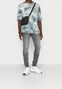 T-shirt gris tie-dye, jeans ajustés gris clair, sac bandoulière noir avec logo, et baskets blanches avec accents gris.