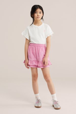 Ein junges Mädchen steht und trägt ein weißes Oberteil mit Rüschenärmeln, einen rosa Rüschenrock, weiße Socken und silberne Schuhe mit pinken Schnürsenkeln vor neutralem Hintergrund.