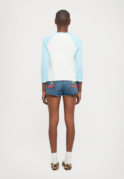 Person with short hair standing facing away wearing a white and light blue shirt, denim shorts with cherry patches, white socks, and leopard slippers.
