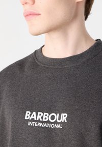 Tmavě šedá mikina s kulatým výstřihem vyrobená z měkké látky, s bílým potiskem textu "BARBOUR INTERNATIONAL" na přední straně.