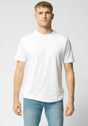 Noor mees seisab näoga otse, seljas tavaline valge lühikeste varrukatega T-särk ja helesinised teksad, taust on ühevärviline.