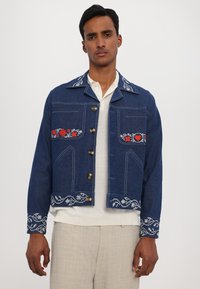 Modra denim jakna z kontrastnim belim vezenjem, ki vključuje rdeče cvetlične vzorce na žepih na prsih in okrasni zaključek na manšetah.