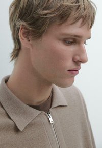 Beiger Strickpullover mit Reißverschluss und spitzem Kragen, getragen über einem braunen Hemd, zeichnet sich durch eine glatte Textur und ein klassisches, tailliertes Design aus.