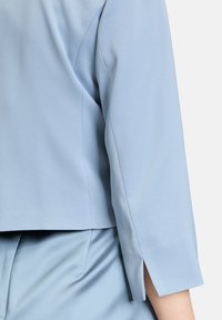 Jasnoniebieska dopasowana marynarka o gładkiej fakturze, z rękawami do łokcia i rozcięciami na bokach u dołu, zapewniająca dopasowany kroj.