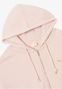Veste à capuche zippée rose clair avec une fermeture éclair dorée et un cordon de serrage. Elle présente une capuche et un matériau intérieur doux, avec un branding minimal sur la poitrine.
