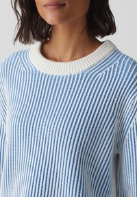 Kvinna som bär en blå och vit vertikalt randig stickad tröja med en tjock vit ribbad rund halsringning.