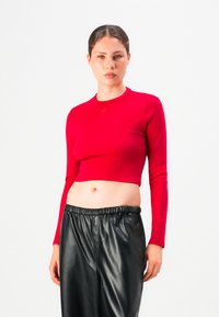 Woman wearing a fitted red long-sleeve crop top and loose black faux leather pants against a plain white background.