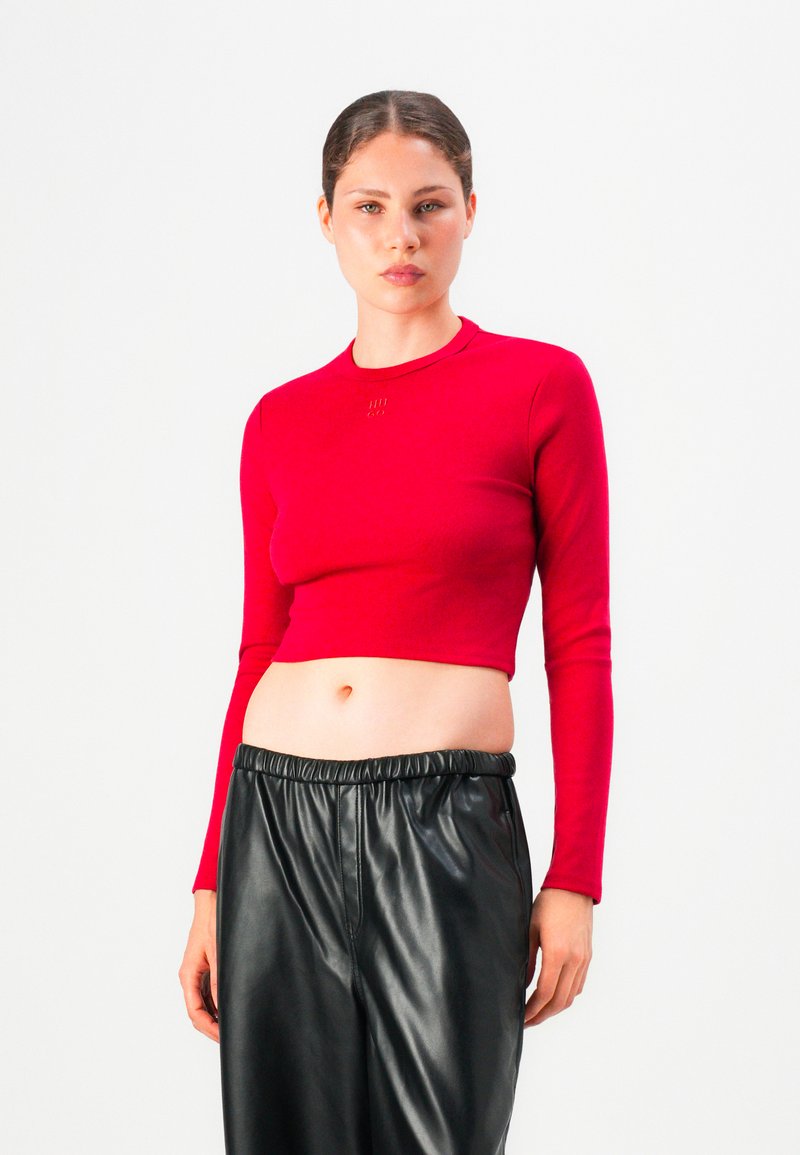 Woman wearing a fitted red long-sleeve crop top and loose black faux leather pants against a plain white background.