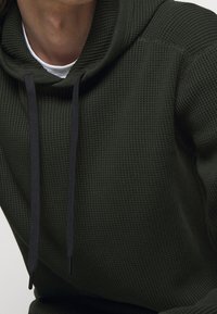 Sudadera con capucha de color verde oscuro con un patrón de gofre texturizado. Presenta cordones negros y una capa interior blanca en contraste.
