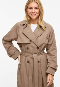Trench-coat beige avec un design à double boutonnage, de grands revers et une ceinture à la taille. Présente des accents de boutons et des manches retroussées.