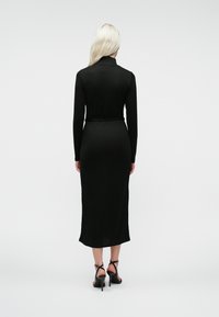 Žena s dlhými blond vlasmi, oblečená v čiernych midi šatách s dlhým rukávom a čiernych sandáloch na vysokom opätku, stojí otočená chrbtom.