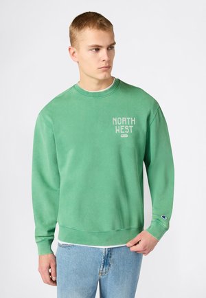 Giovane uomo che indossa una felpa verde con la scritta "NORTH WEST" sul petto e jeans azzurri, guarda verso destra.