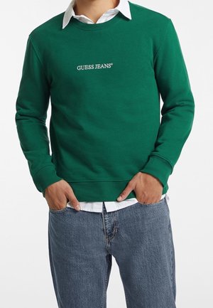 Personne portant un sweat-shirt vert avec le texte « GUESS JEANS » sur une chemise blanche à col et un jean bleu, les mains partiellement dans les poches.