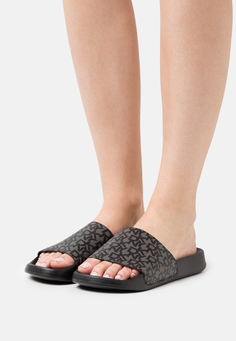 Personne portant des sandales slide noires avec des larges brides grises à motif, pieds nus sur fond blanc.
