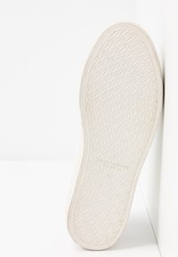 Talpa de sneaker albă cu un model de cauciuc texturat și o ușoară curbură. Prezintă un nume de marcă și detalii despre dimensiune embosate în apropierea călcâiului.