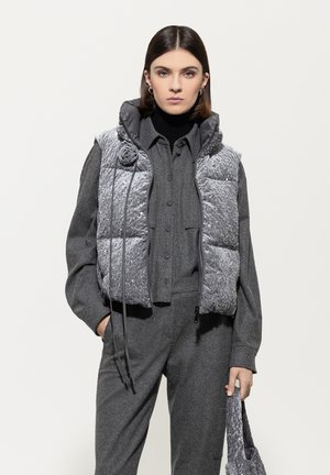 Gepolsterte graue Weste mit strukturierter Oberfläche, mit Kapuze, Frontreißverschluss und dekorativem Rosendetail. Kombiniert mit einem grauen Outfit.