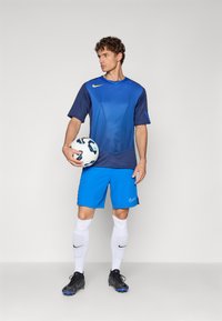 Tricou sportiv albastru cu un design texturat, asortat cu pantaloni scurți albaștri. Ținând o minge de fotbal, poartă șosete albe și ghete negre.