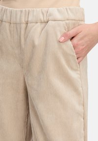Béžové kordové kalhoty s vertikálním žebrovaným vzorem, s elastickým pasem a bočními kapsami, na obrázku je ruka odpočívající na pravé kapse.