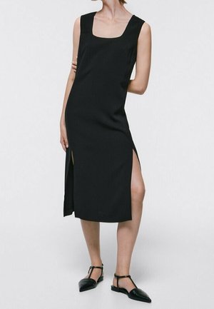 Woman wearing a sleeveless black midi dress with side slits, paired with black T-strap pointed flats, standing against a white background.