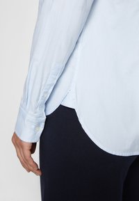 Camicia button-up azzurro chiaro con maniche lunghe, caratterizzata da un orlo curvo e polsini e una texture liscia. Indossata sopra un tessuto scuro e testurizzato.