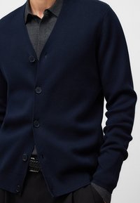 Marineblauer Cardigan aus Strickmaterial, mit V-Ausschnitt, fünf Knöpfen und geripptem Saum. Tragen über einem dunkelgrauen Hemd mit Kragen.