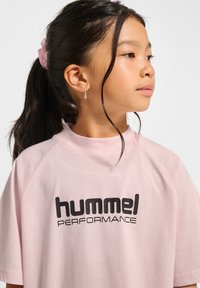 Różowa koszulka z krótkim rękawem i okrągłym dekoltem, z czarnym logo "hummel PERFORMANCE" umieszczonym na środku z przodu w wyraźnym kroju.