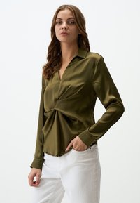 Blouse olive, à manches longues, avec col et détail en torsion à l'avant, en tissu doux. Associée à un pantalon blanc, coupe slim.