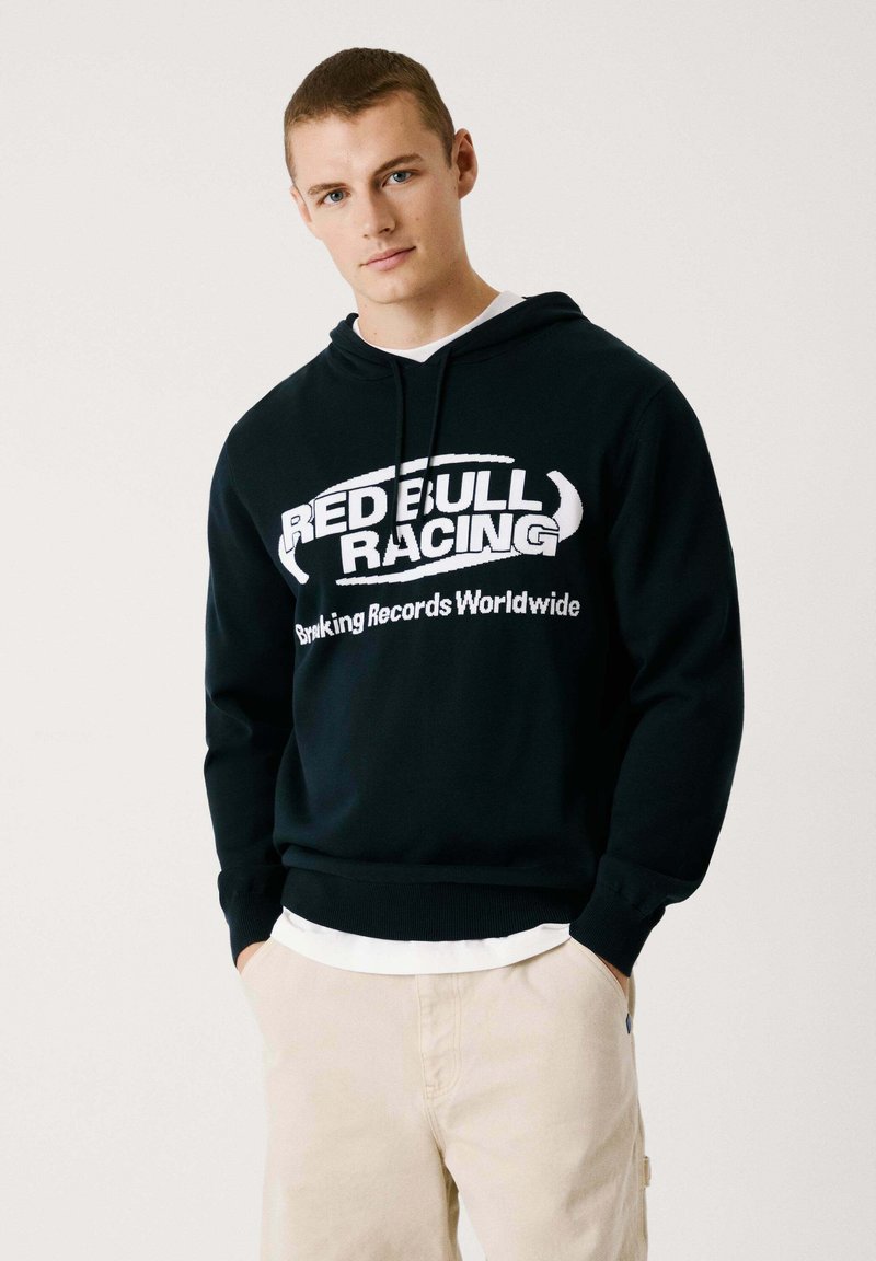 Svart hoodie med en vit "RED BULL RACING" logotyp och texten "Ranking Records Worldwide." Har en justerbar huva och ribbade ärmslut.