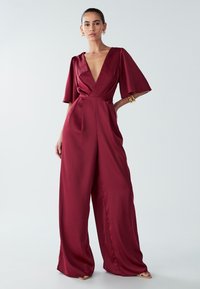 Mujer vistiendo un mono de satén rojo profundo con piernas anchas y mangas flotantes, de pie con una mano en la cadera frente a un fondo sencillo.