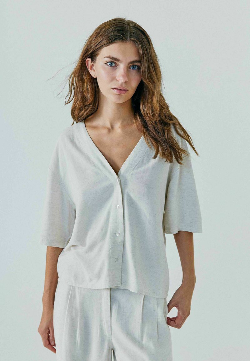 Jeune femme aux longs cheveux bruns et aux yeux bleus, portant une chemise ample claire boutonnée et un pantalon assorti sur un fond uni.