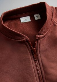 Detailný záber na hrdzavú detskú bundu s plastovým zipsom a štítkom s označením veľkosti pre vek 6 rokov.