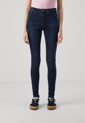 Donkerblauwe slim-fit jeans gedragen met zwart-witte sneakers en witte sokken, getoond van taille tot voeten bij een persoon met een roze top.