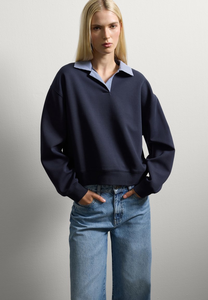 Mujer con cabello rubio largo, vistiendo un suéter azul marino con una camisa de cuello azul debajo y jeans de color azul claro, con las manos en los bolsillos.