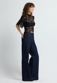 Top in pizzo nero con maniche corte, colletto alto e design corto abbinato a jeans a vita alta di colore blu scuro a gamba larga con dettagli dorati.