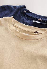 Deux t-shirts en coton à col rond pliés en beige et bleu marine, empilés sur une surface blanche.