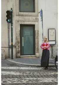 Różowy i czerwony sweter w romby, czarna długa spódnica, stojąca przy zielonym sygnale dla pieszych, na tle fakturowanej kamiennej ściany i szklanych drzwi.